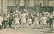 De Mariaschool Pastoor van Erpstraat 4. Kinderen op de speelplaats met bloemenkrans en in het wit en kinderen in hun dagelijkse kleren. Voor meer details klik hier.